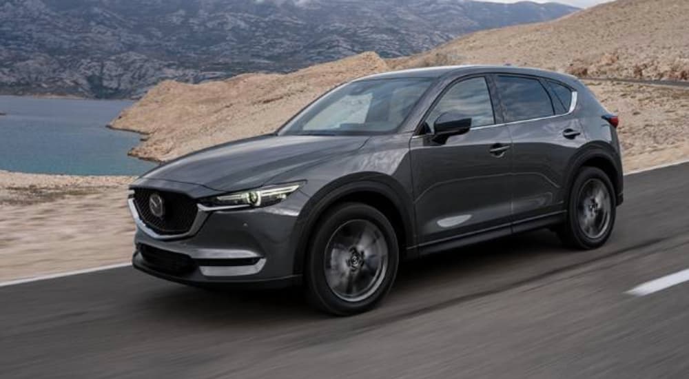 A grey 2020 Mazda CX-5 is driving past a lake and mountain.