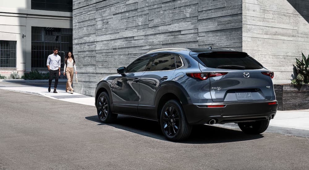 A couple is walking towards a gray 2021 Mazda CX-30, shown from the rear, that is parked in front of a stone wall.