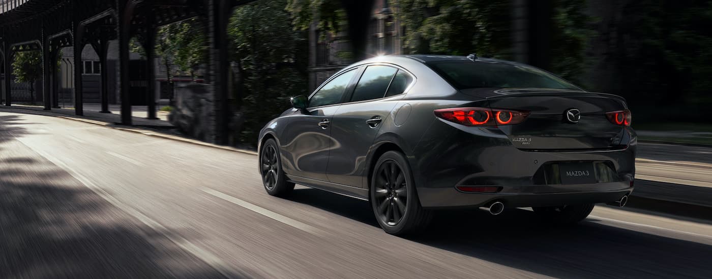 A grey 2021 Mazda 3 sedan is shown from the rear driving down an empty street.