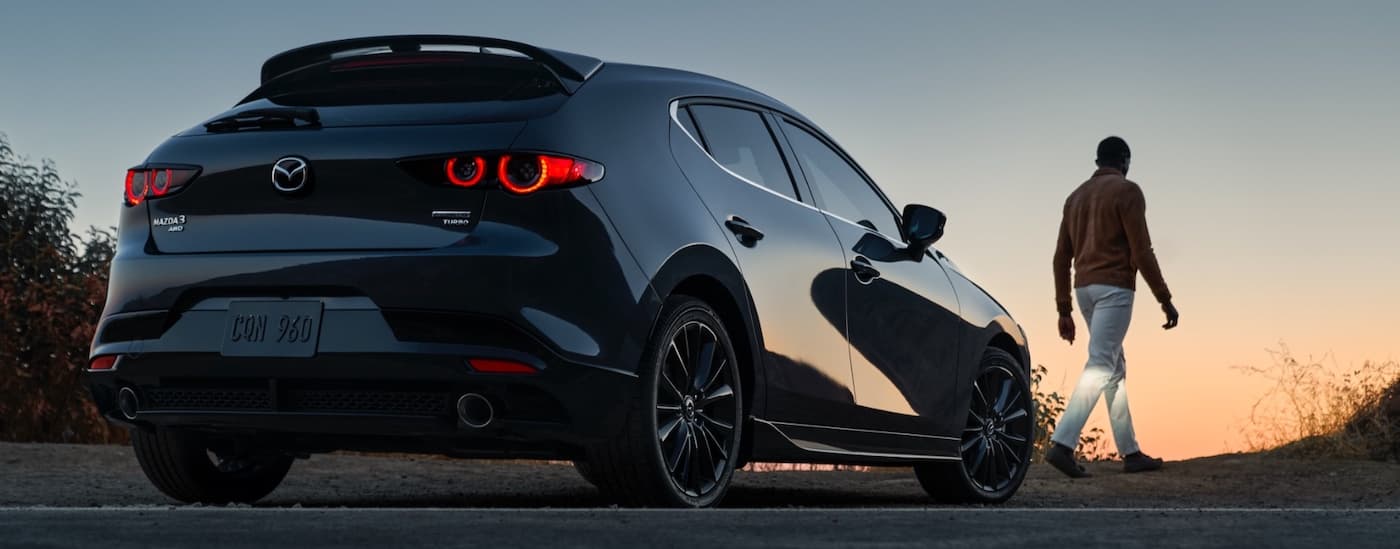 A grey 2021 Mazda3 hatchback is shown from the rear next to a person at dusk after leaving a Mazda3 dealer in Houston.